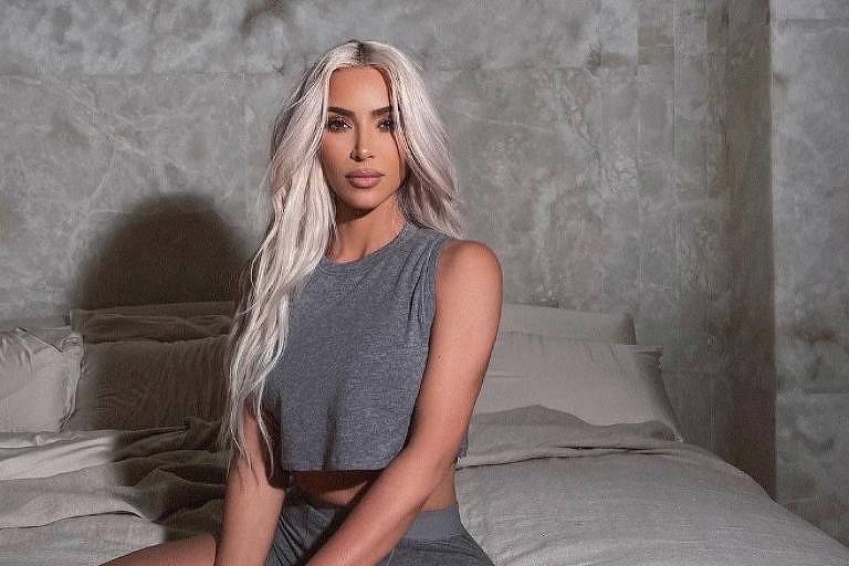 Em foto colorida, Kim Kardashian, de conjunto cinza, aparece sentada em cima de uma cama de casal