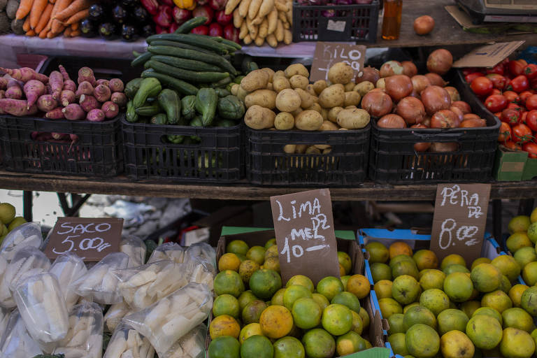 Imagem mostra caixotes com legumes e frutas em uma feira.