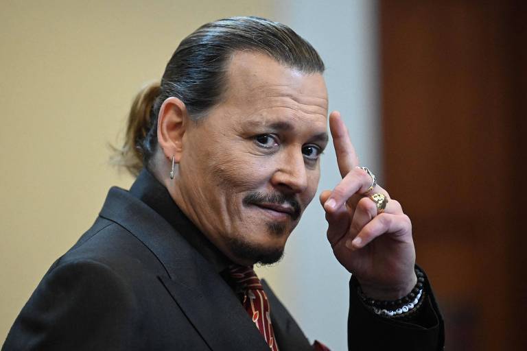 Johnny Depp, de terno e gravata, sorri para as câmeras, com o dedo indicador apoiado na testa