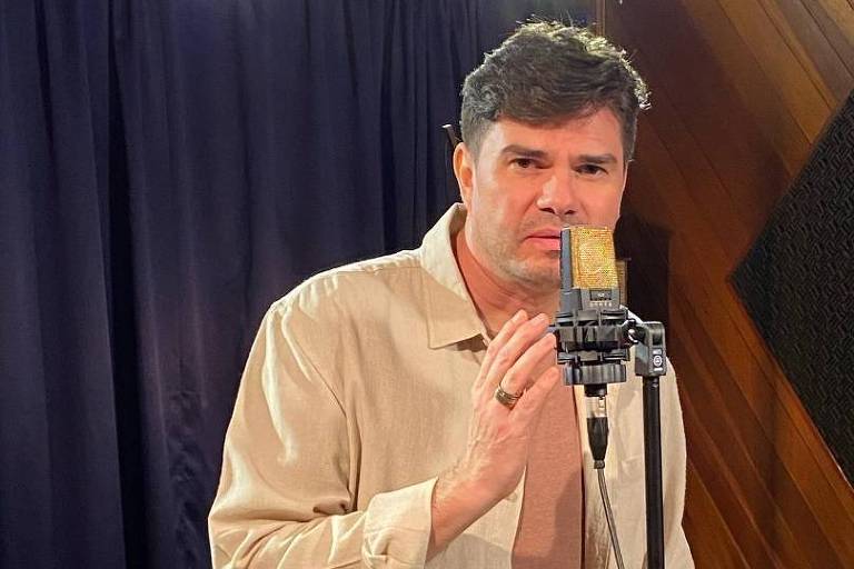 Em foto colorida, homem branco vestido de camisa bege de mangas compridas aparece cantando em um estúdio e posa em frente ao microfone