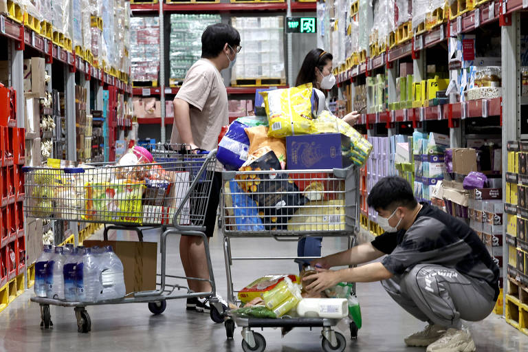 Imagem mostra três pessoas num corredor de um supermercado, em meio a duas estantes com produtos. Eles estão juntas a dois carrinhos cheios de compras.