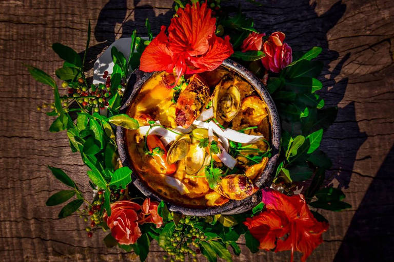 A foto mostra uma panela de barro com um ensopado de frutos do mar. A panela aparece envolta em uma coroa de folhas e flores, sobre uma mesa de madeira rústica