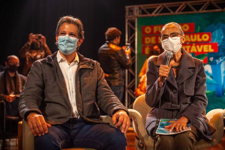 Fernando Haddad e Marina estão ambos sentados, um lado do outro, os dois usam máscaras faciais e ela segura um microfone