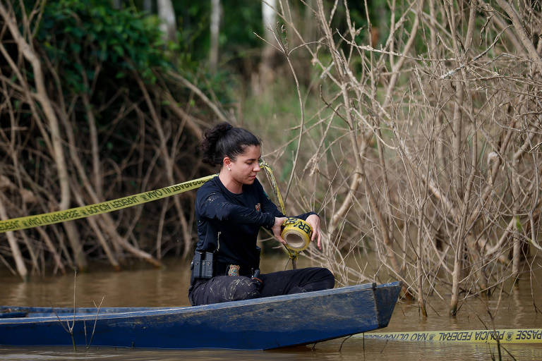 Mulher policial em canoa passa fita amarela para isolar área de crime na margem de rio