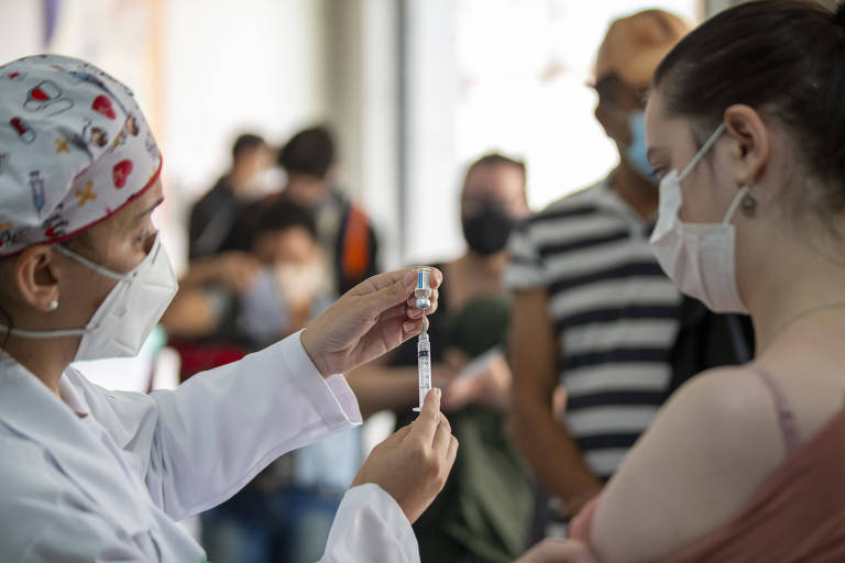 Profissional da saúde mostra dose de vacina para pessoa. As duas usam máscara 