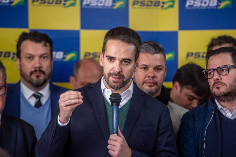 Político fala ao microfone cercado de apoiadores. Há pano de fundo atrás, azul e amarelo, com a marca do PSDB