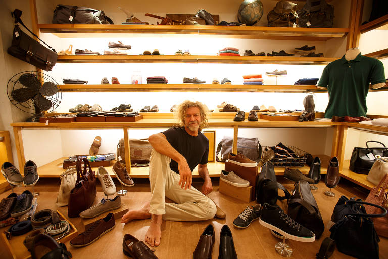Imagem mostra homem sentado em chão de madeira de uma loja, no meio de vários sapatos de couro, camisas e outros itens de vestuário.