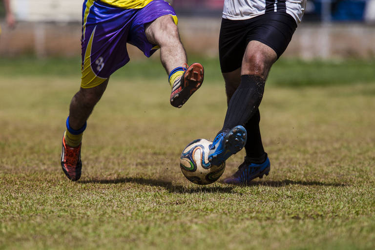 Dois jogadores, os quais somente as pernas aparecem na imagem, participam de jogo de futebol de várzea em São Paulo