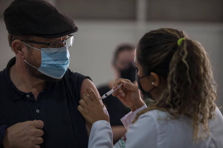 Mulher de rabo de cavalo e avental branco aplica vacina em homem de chapéu e camisa pretos