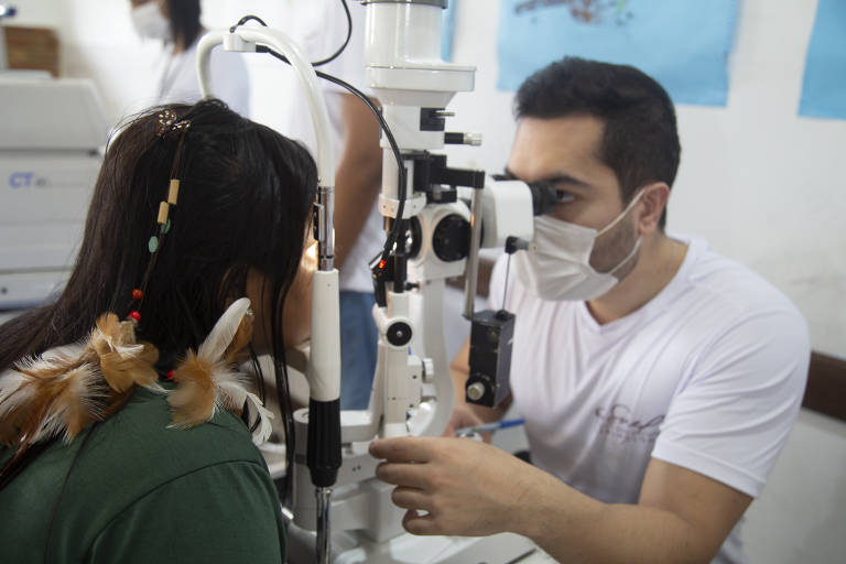 Na foto estão duas pessoas, uma mulher que aparece em frente a um aparelho de exame oftalmológico. Do outro lá do aparelho está um homem que opera a máquina
