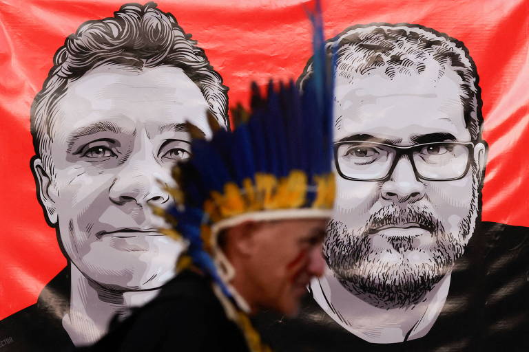 Indígena com cocar de perfil direito com painel vermelho com imagens dos rostos de Dom Phillips e Bruno Pereira em preto e branco