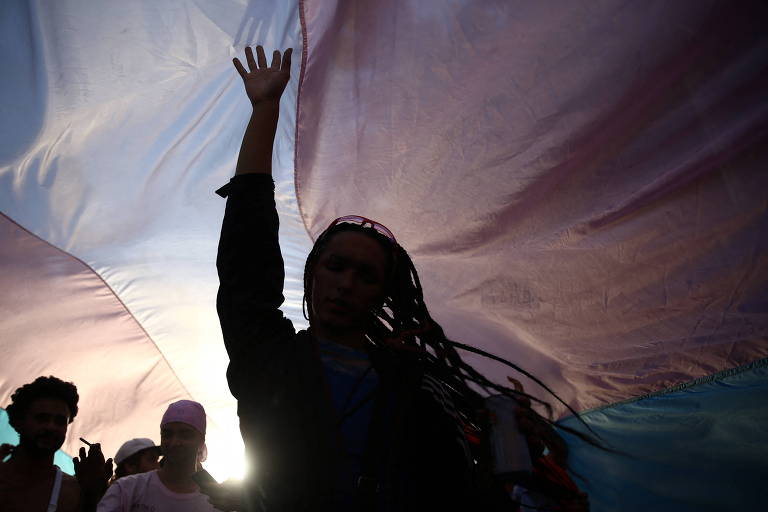 manifestantes vistos sob grande bandeira com as cores LGBT