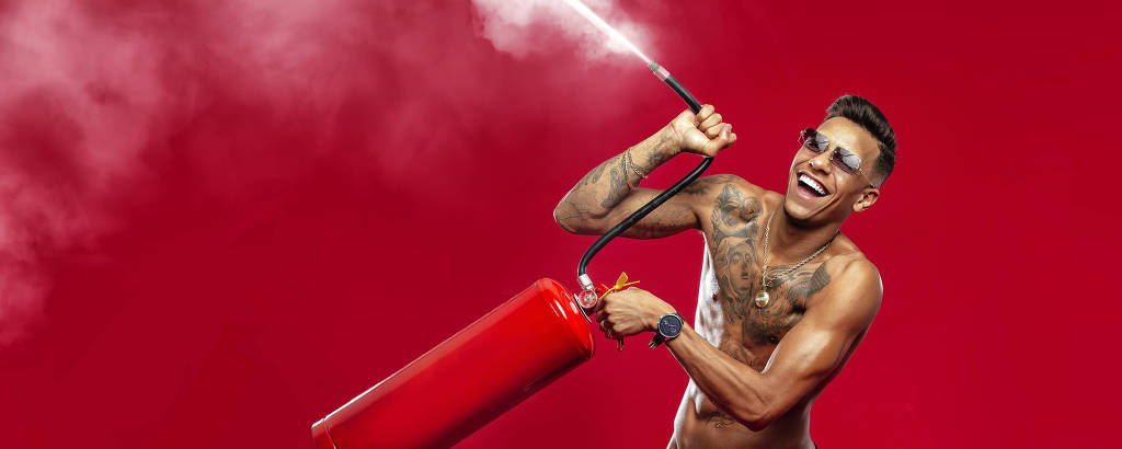 homem moreno e tatuado posa sem camisa segurando extintor de incêndio diante de fundo vermelho