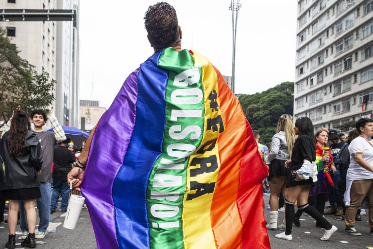Imagem mostra homem de costas carregando bandeira LGBT, na qual está escrito "Fora Bolsonaro". Ele está na rua, em meio a outras pessoas.