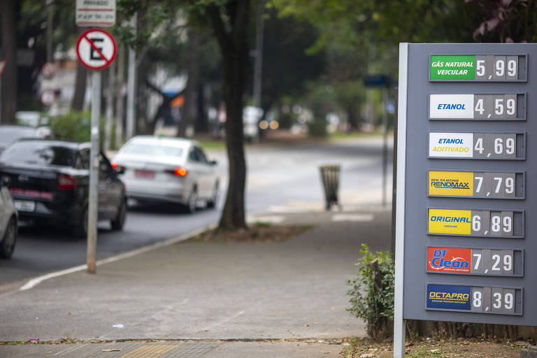 À direita, painel cinza com preços de diferentes combustíveis. À esquerda, calçada e rua com carros