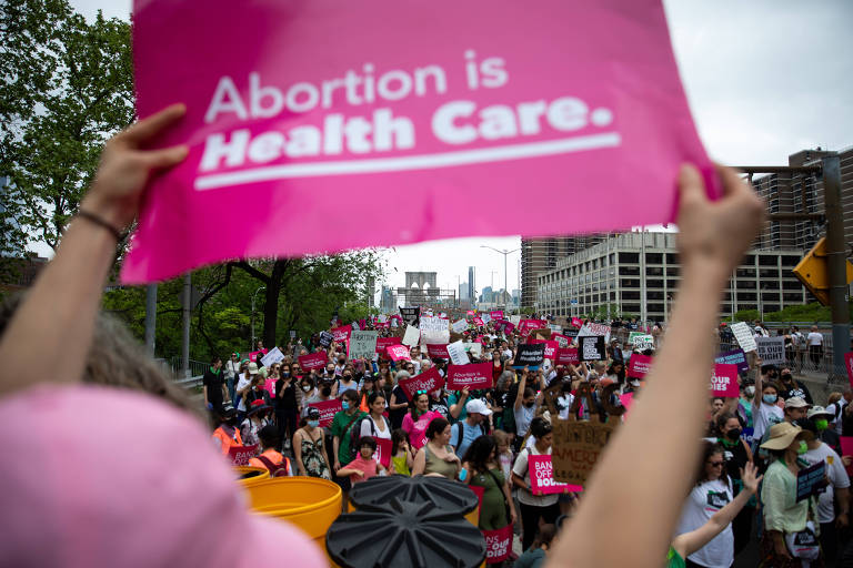 Imagem mostra manifestante erguendo cartaz rosa com a frase "Abortion is Health Care" em branco. Ela está em primeiro plano, enquanto várias pessoas, com outros cartazes rosa, aparecem ao fundo, onde também é possível ver prédios e árvores.