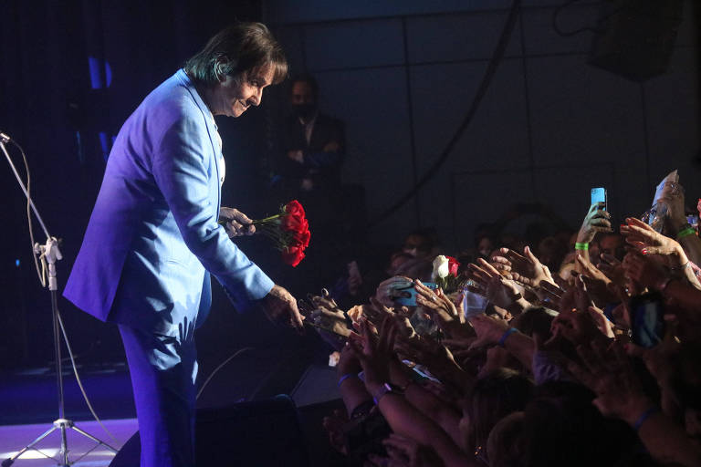 Em foto colorida, homem de terno azul distribui rosas vermelhas do palco para a plateia de um show