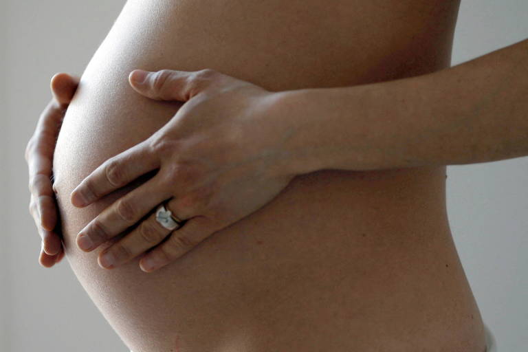 Uma mulher branca grávida está com as mãos sobre a barriga