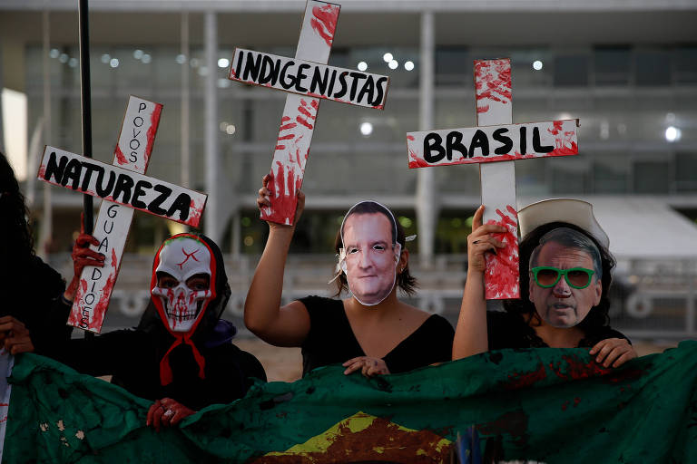 Pessoas usam máscaras de caveira e com as caras de Bolsonaro e Xavier e seguram cruzes manchadas de tinta vermelha