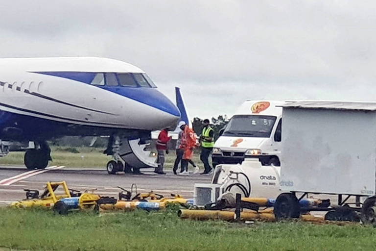 Neymar, em uma jaqueta laranja, caminha de uma van em direção a uma aeronave azul e branca escoltado por outras pessoas. A foto é de longe e mostra um gramado e parte da pista do Aeroporto Internacional de Boa Vista.