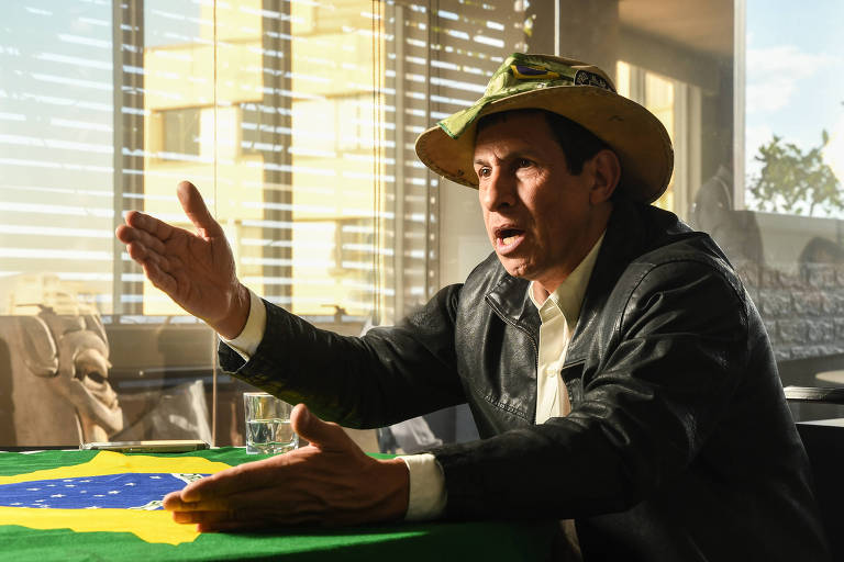 Homem veste jaqueta, e chapéu, está com gesticulando com as mãos e pela expressão facial parece estar falando de modo exaltado. À sua frente, na mesa, está estendida uma bandeira do Brasil