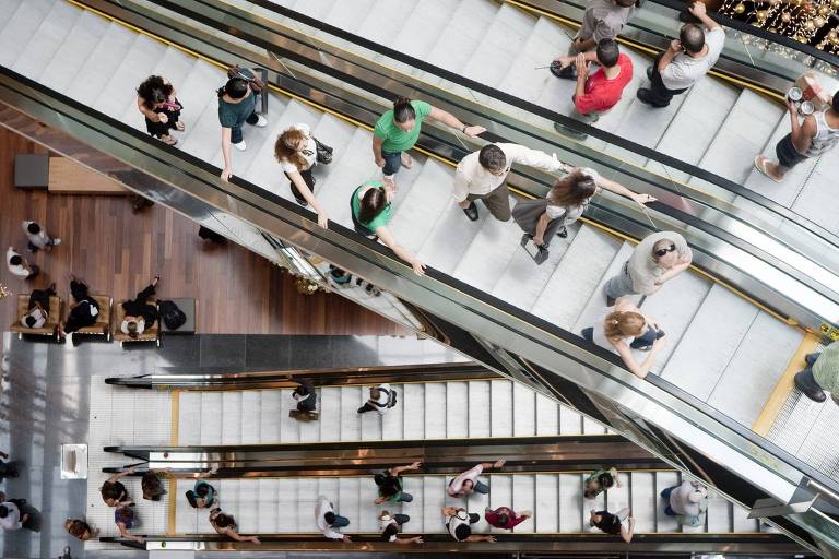 Imagem mostra escadas rolantes de shoppings, com pessoas nelas. A foto foi tirada de cima.