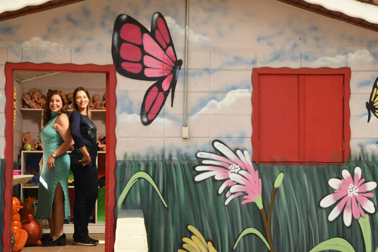 Mulher de vestido verde e outra de blusa e calça pretas estão de costas uma para a outra dentro de uma casinha colorida para crianças, com porta vermelha e borboletas pintadas voando