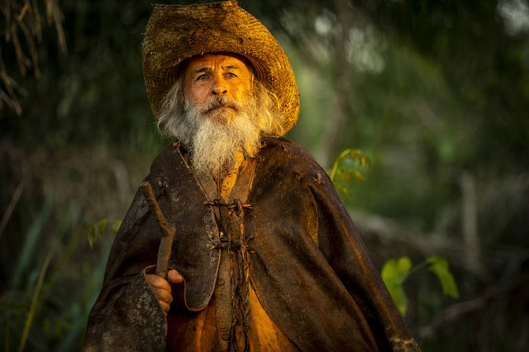 Em foto colorida, homem barbudo de chapéu e cajado aparece no meio da floresta