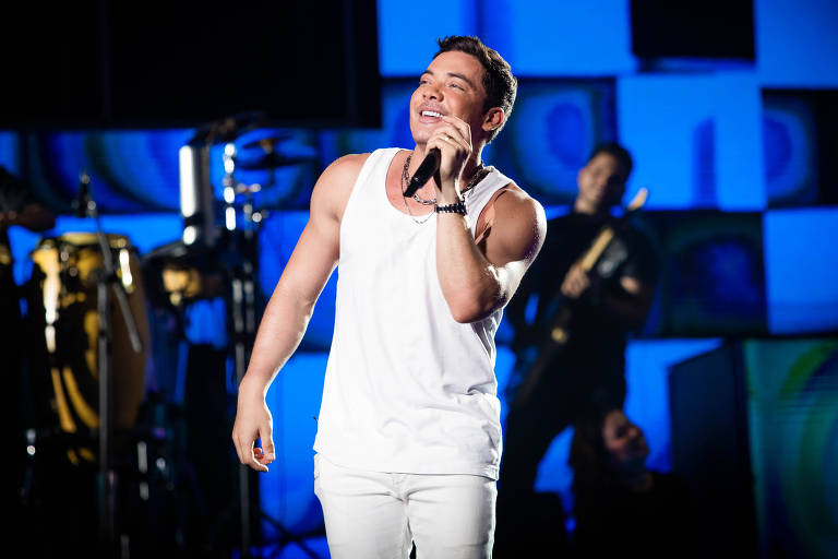 Em foto colorida, homem de calça e blusa branca segura microfone em cima do palco e sorri para o fotógrafo