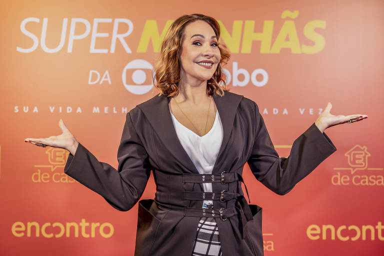 Maria Beltrão sorri e abre os braços posando para foto; ela veste uma blusa branca e um casaco escuro