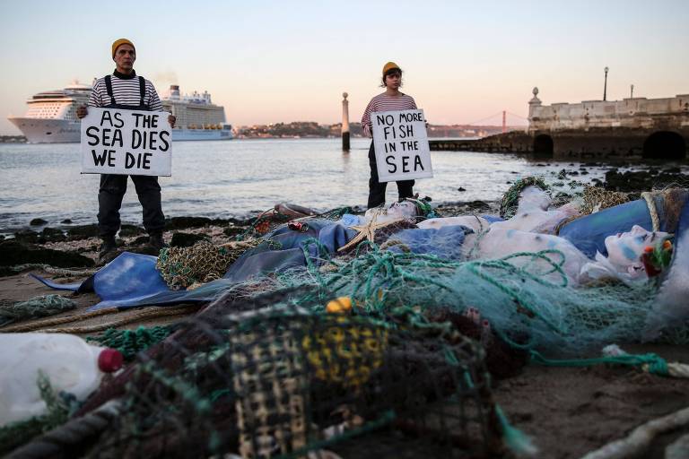 Duas pessoas seguram cartazes com os dizeres  "As the sea dies we die" e "No more fish in the sea"; no chão outras pessoas, fantasiadas como peixes, estão presas em redes e pedaços de lixo