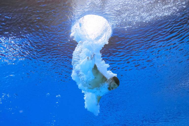 Visto de dentro da piscina, atleta adentra água, criando bolhas de ar em torno de seu corpo