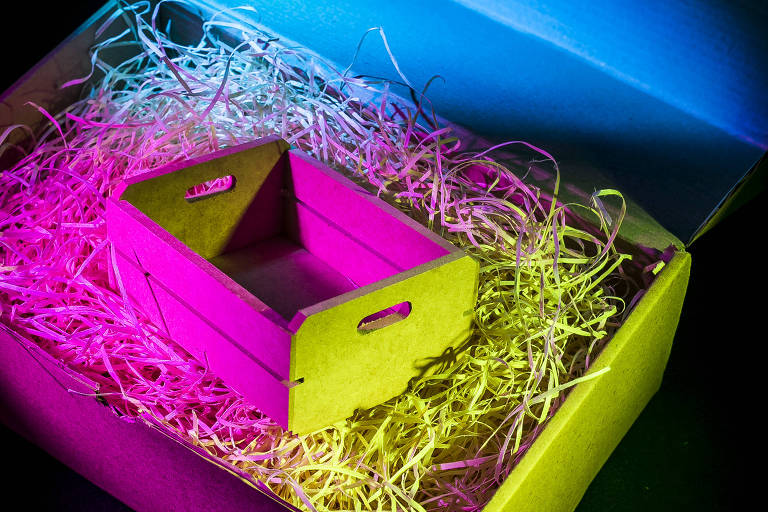 Fotografia mostra uma caixa de madeira cercada por palha dentro de uma caixa de papelão; os objetos estão iluminados com luzes nas cores rosa e azul
