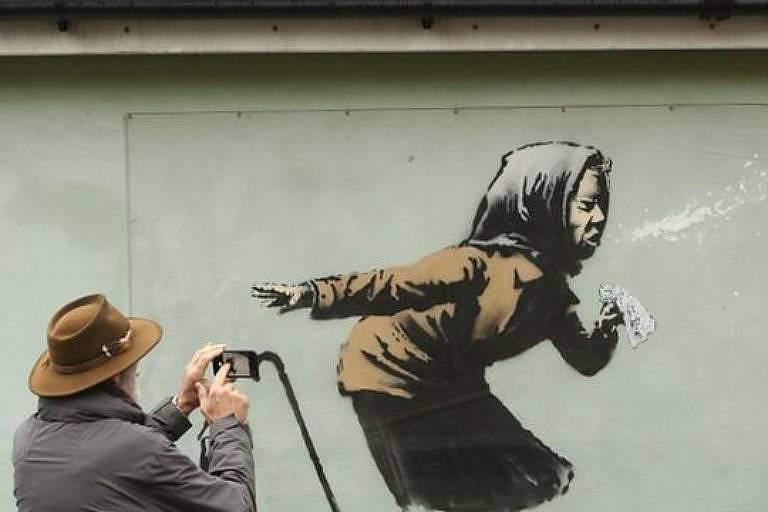 Imagem mostra homem com selular na mão fotografando um grafite em uma parede que mostra uma pessoa espirrando