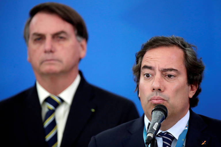 Pedro Guimarães aparece na direita discursando em frente a um microfone. Bolsonaro está do lado esquerdo, mais ao fundo.