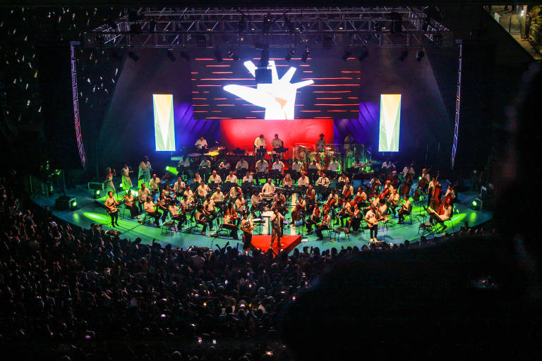 Foto da apresentação da orquestra sinfônica. A banda está no centro do palco, por sua vez iluminado em verde e azul, enquanto uma multidão lota a plateia.