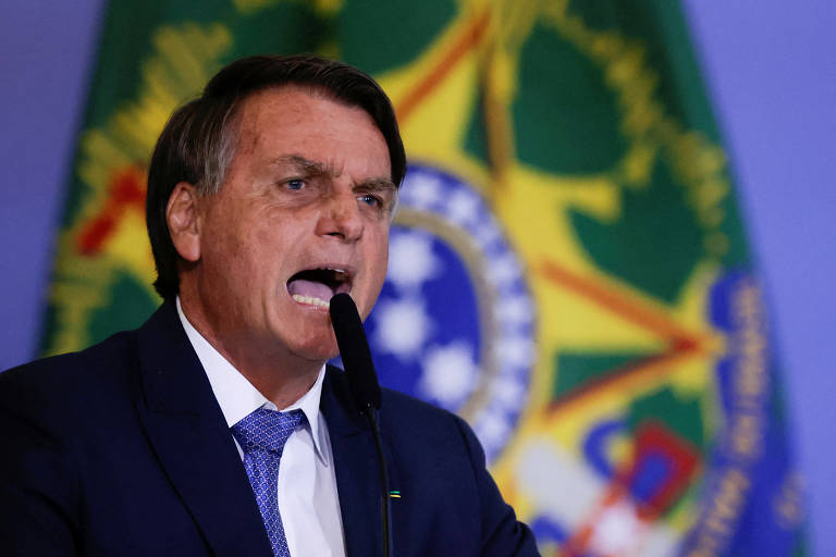 Na foto, o presidente Jair Bolsonaro. Ele é um homem branco, com cabelos um pouco grisalhos e pele mais avermelhada, com manchas. Ele aparece com uma cara de bravo, como se estivesse gritando. Veste um terno preto, gravata azul e camisa social branca. Ao fundo, a bandeira da República