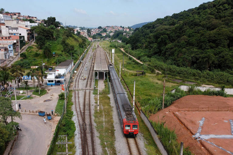 Imagem mostra trem em frente à estação. Ao lado direito, há um morro cheio de vegetação. No lado oposto, há uma comunidade com casas de tijolos.