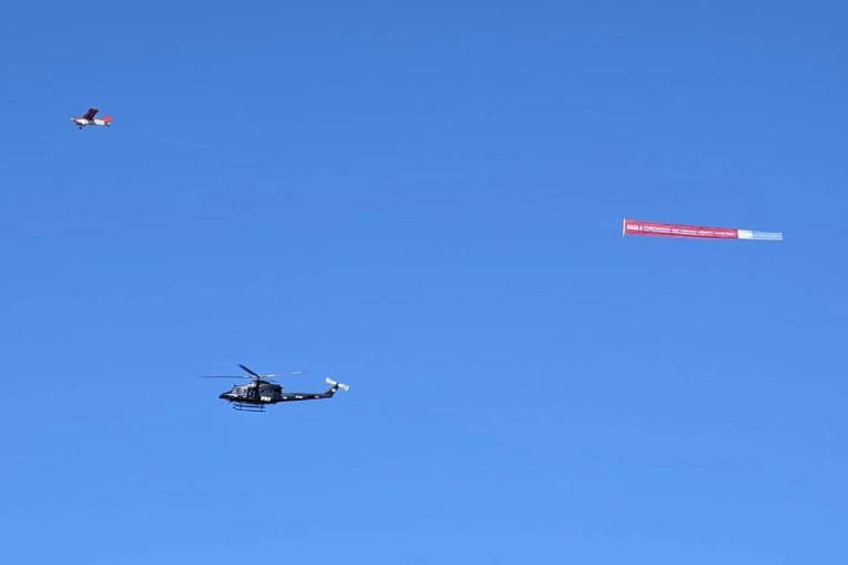 Sob céu azul, avião vermelho de pequeno porte puxa uma faixa vermelha em que se lê em branco os dizeres: 'Nada a comemorar: Bolsonaro mentiu para os PRFs’. Abaixo da faixa, há um helicóptero.
