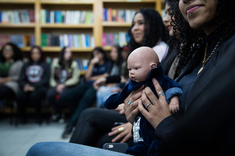 Imagem destaca uma menina segurando um bebê de brinquedo no colo; ela está em uma sala de aula, sentada em uma roda com outros alunos