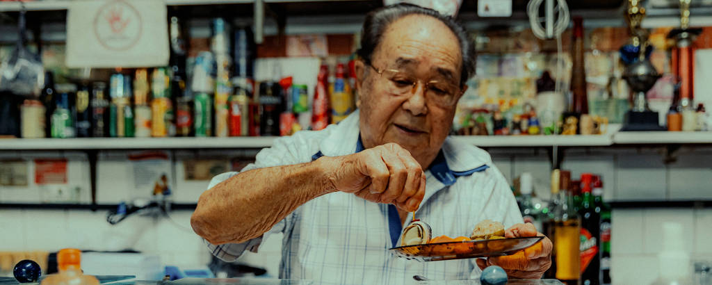 Homem idoso monta prato com itens frios em frente a prateleira de bebidas em boteco