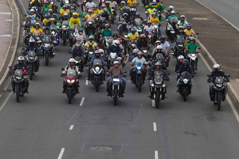 Vários motoqueiros ocupam mais da metade de uma pista de uma avenida. Ao centro, a frente do grupo, o presidente Bolsonaro, que veste jaqueta marrom e capacete branco. Vários motoqueiros vestem amarelo e verde e levam bandeiras