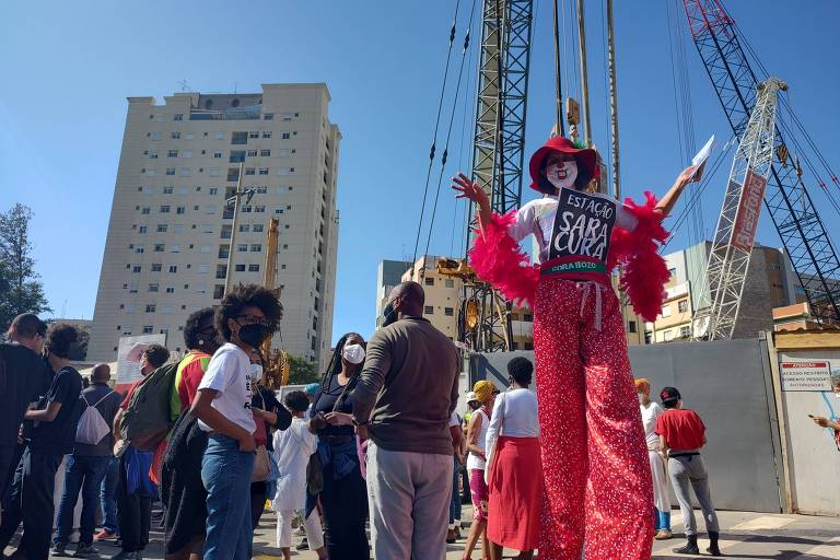 Mulher em perna de pau, com toupa vermelha, chapéu e máscara, defende a mudança de nome da futura estação 14 Bis