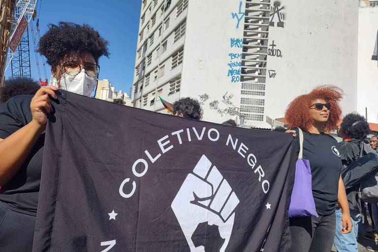 Duas garotas negras, usando roupa preta e máscaras, seguram bandeira onde lê-se: Coletivo Negro