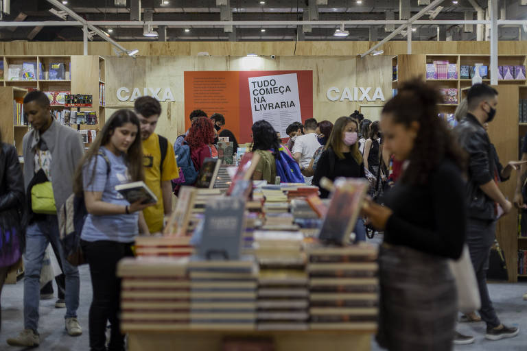 A imagem mostra uma feira de livros com várias pessoas circulando entre estantes. No primeiro plano, há uma mesa com livros empilhados. Algumas pessoas estão lendo ou segurando livros, enquanto outras conversam. Ao fundo, há estantes de livros e uma parede com uma faixa que diz 'Todo livro é uma viagem'.