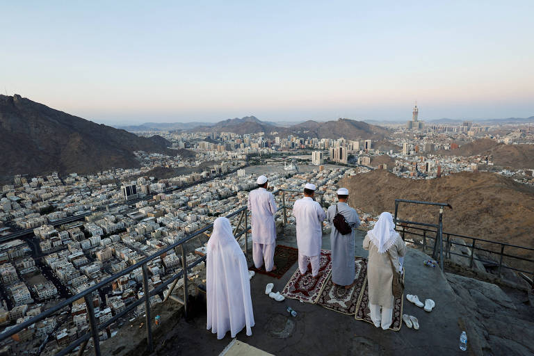 Cinco muçulmanos fazem oração em tapetes sobre cume de monte, com visão da cidade de Meca ao fundo