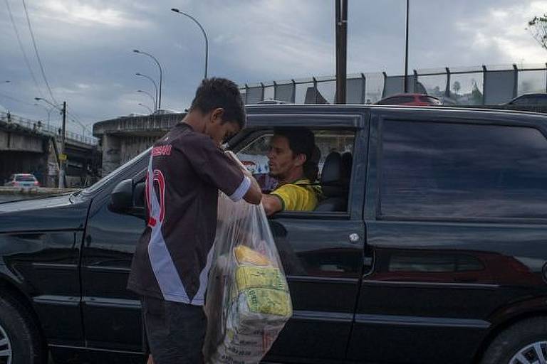 Imagem em primeiro plano mostra uma criança com um saco cheio de biscoito Globo ao lado de um carro parado. O motorista está com o vidro aberto.