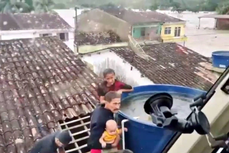 imagem de trecho de vídeo de bebe no colo de bombeiro que se apoia em caixa dágua em escada em um telhado