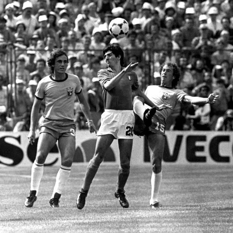 Foto em preto e branco. Paolo Rossi tenta cabecear a bola em Brasil versus Itália na Copa do Mundo da Espanha. Ele é observado por Oscar, que está à esquerda, e marcado por Falcão, que está a direita