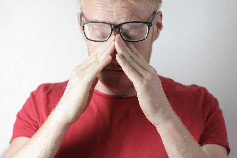 Homem grisalho, branco, de óculos e camiseta vermelha, massageando a ponte do nariz